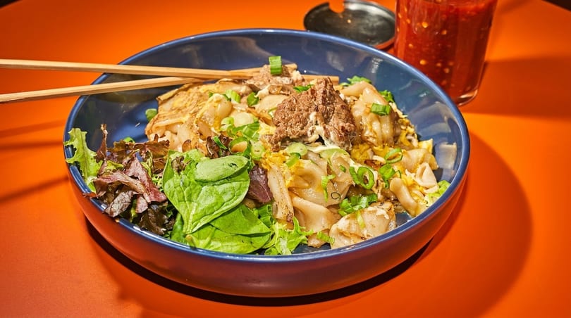Noodle dish with beef, egg, green onions, and lettuce in a blue bowl with chopsticks.